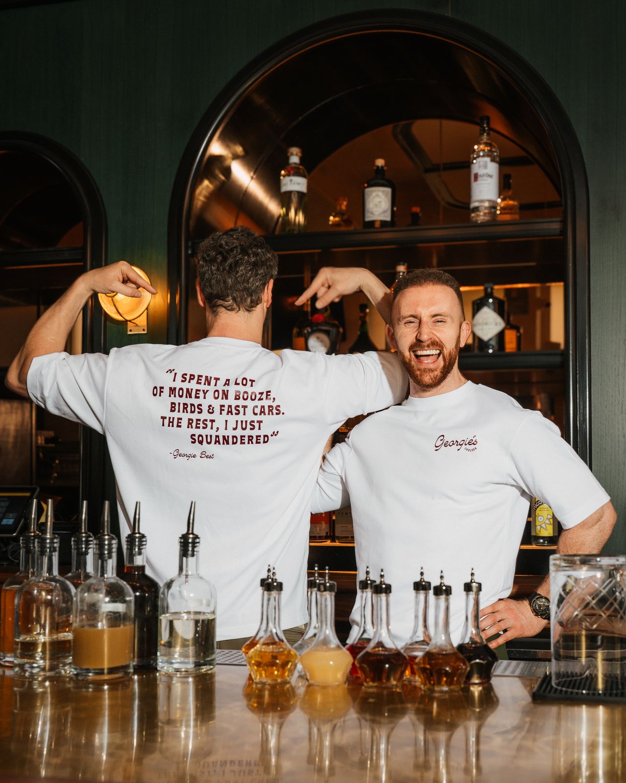 Two Georgie's bartenders laughing behind the bar, the arched back-bar mirror and bottles behind them.